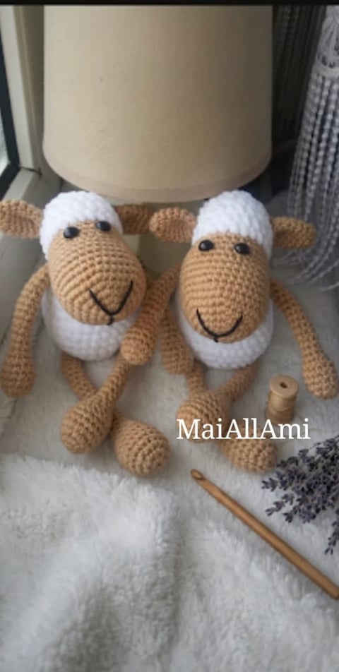 Two crocheted sheep amigurumi toys with tan bodies and white wool heads, smiling faces, standing under a cream-colored lamp on white fabric