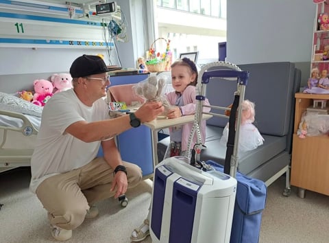 Adult man crouching while playing with a young child in a hospital room surrounded by luggage and stuffed animals