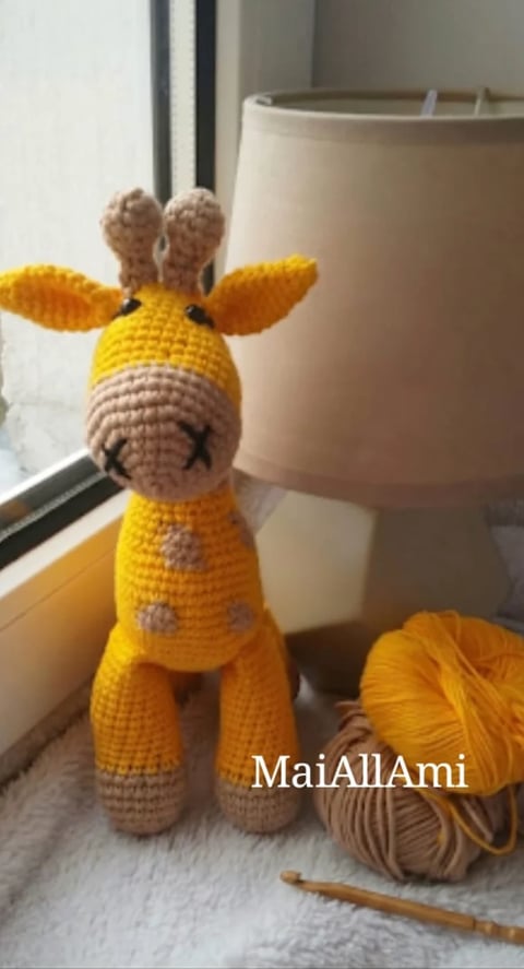 Handmade yellow and tan crocheted giraffe toy with a smiling face, displayed next to yarn balls and a crochet hook on a white surface