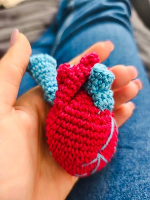 Small crocheted red and blue heart with wings held in a hand over someone wearing blue jeans