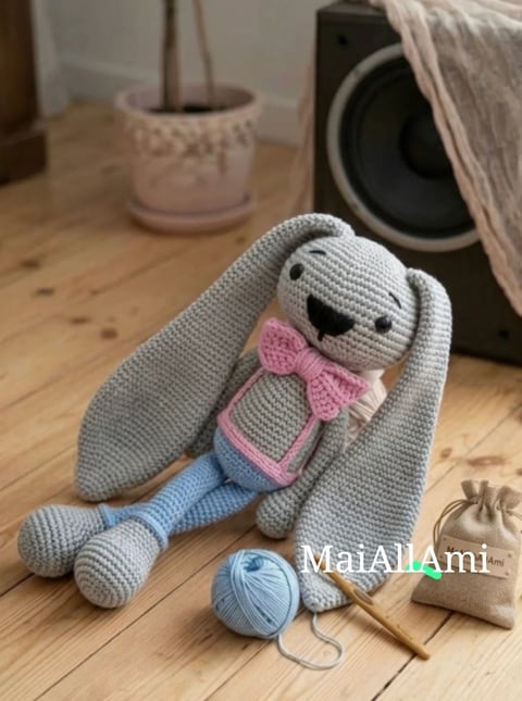 Crocheted gray rabbit with pink bow and blue pants sitting on wooden floor with yarn and crochet hook nearby