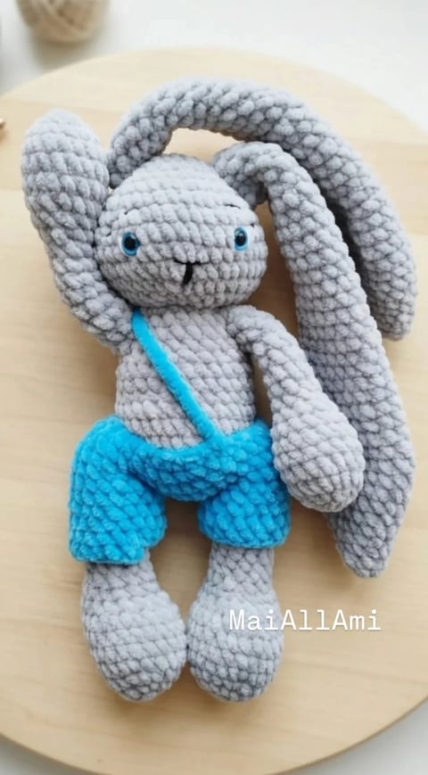 Gray crocheted bunny amigurumi with long ears and turquoise shorts sitting on a cream-colored plate
