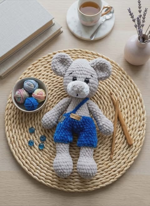 Crocheted gray teddy bear wearing blue shorts on a woven placemat surrounded by craft supplies and coffee