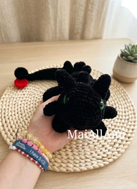 Hand holding black crocheted cat plushie with green eyes on woven placemat, beaded bracelet visible