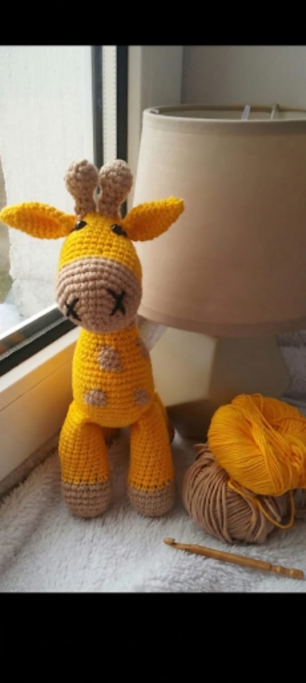 Crocheted yellow giraffe toy with beige spots and ears, displayed on a windowsill with yarn and crochet hook nearby