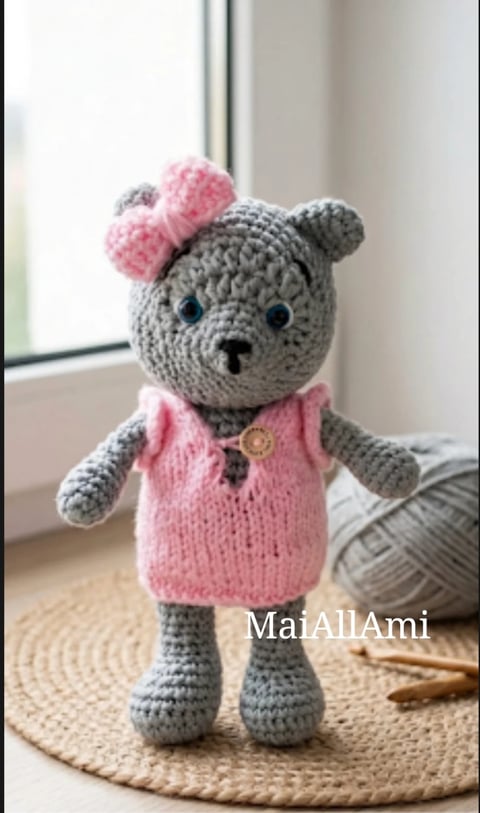 Handmade gray crocheted teddy bear with pink ears, wearing a pink sweater with button, sitting on a woven mat by a window
