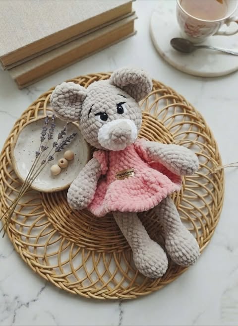 Crocheted gray mouse wearing a pink dress sits on a woven circular placemat with decorative items and a cup of tea