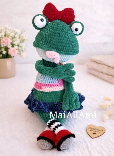 Handmade crochet frog amigurumi doll with red bow, green body, pink stripes, and red shoes on display