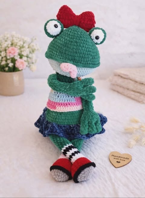 Handmade crochet frog amigurumi with red bow, green body, striped dress, and red shoes on white surface with flowers