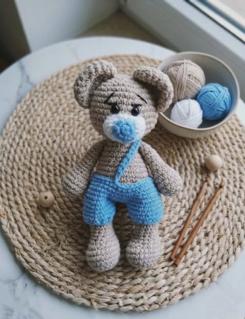 Crocheted amigurumi teddy bear with blue nose and shorts sitting on a beige mat beside yarn and crochet hooks