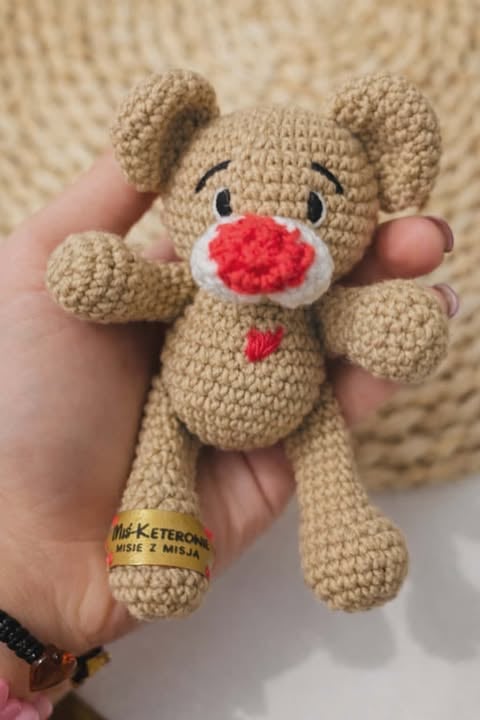 Crocheted tan teddy bear with red nose and embroidered eyes, held in someones hand against a knitted background