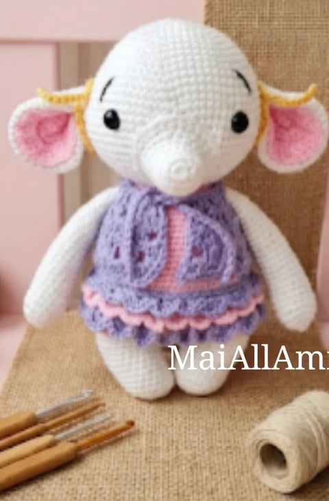 Handmade crochet elephant plushie with white body, pink ears, purple dress, and gold accents, displayed on beige fabric with crafting supplies nearby