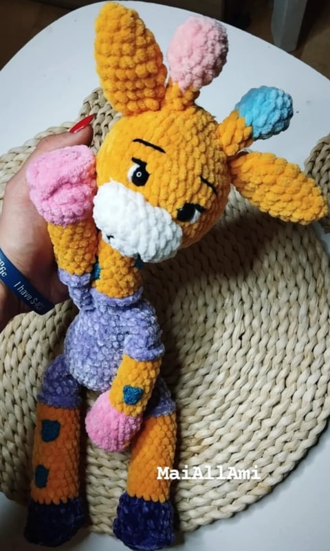 Colorful crocheted giraffe toy with yellow body, pink ears, blue accents, and patterned legs held in someones hand