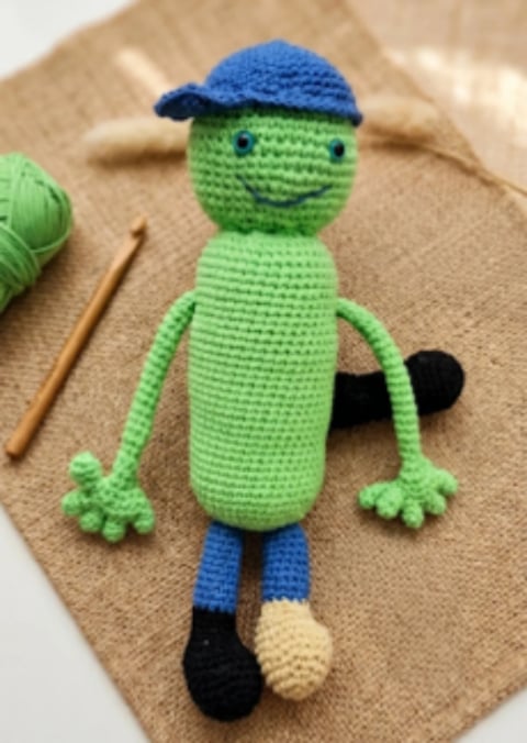 Handmade crocheted green stuffed toy character wearing a blue cap and blue pants with black shoes, displayed on beige fabric