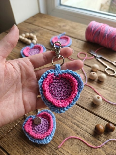 Hand holding crocheted pink and blue heart-shaped keychains with crafting supplies on wooden table