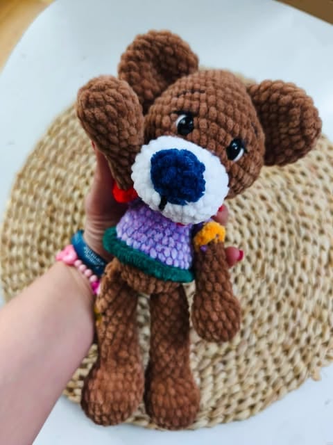 Crocheted brown teddy bear with white muzzle and blue nose, wearing colorful striped sweater, held in hand over beige knitted blanket