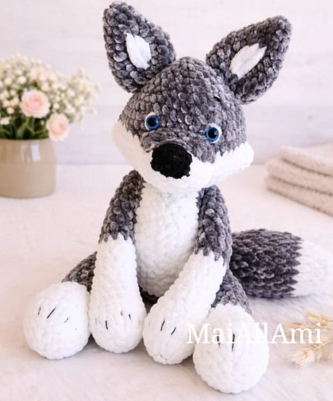 Crocheted amigurumi husky toy with gray and white yarn, blue eyes, sitting on a surface with flowers and towels nearby