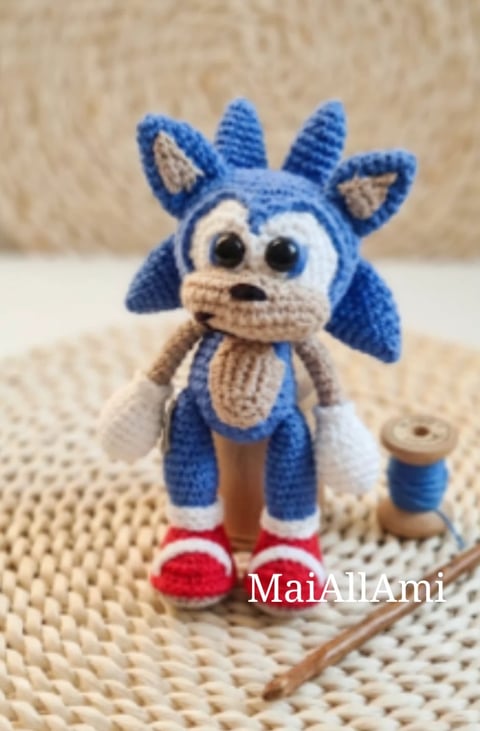 Blue and white crocheted toy character with horns, resembling Sonic the Hedgehog, with red shoes and surprised expression