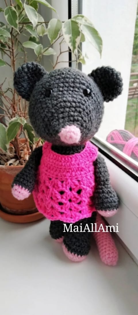 Gray crocheted stuffed bear with pink nose, wearing a hot pink sweater, sitting on a windowsill next to a potted plant