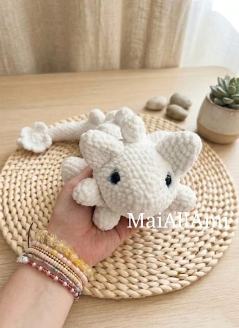 Hand holding a white crocheted elephant amigurumi on a woven placemat with a potted succulent and decorative stones
