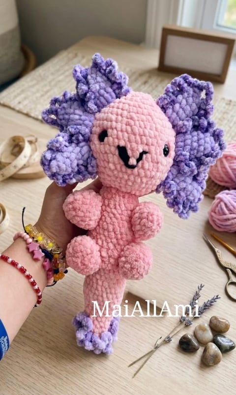 Pink and purple crocheted amigurumi axolotl toy held in a hand on a wooden desk with craft supplies nearby
