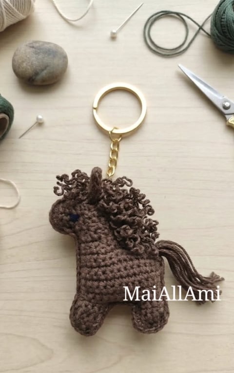 Brown crocheted horse keychain with curly mane and tail on a beige crafting surface surrounded by yarn and tools