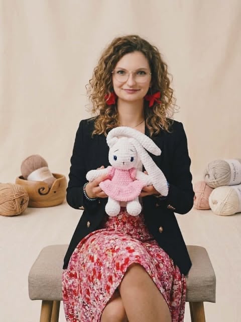 Woman holding a knitted rabbit plush toy, sitting with yarn balls and crafting supplies around her