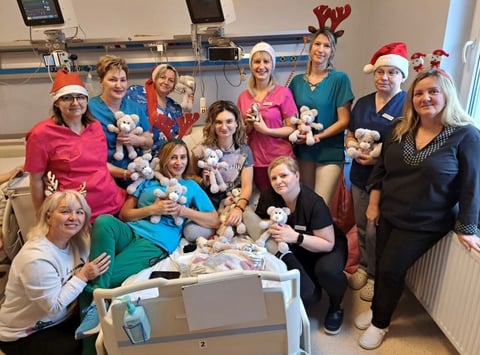 Group of healthcare workers in a clinical setting holding teddy bears and wearing festive hats for a holiday celebration